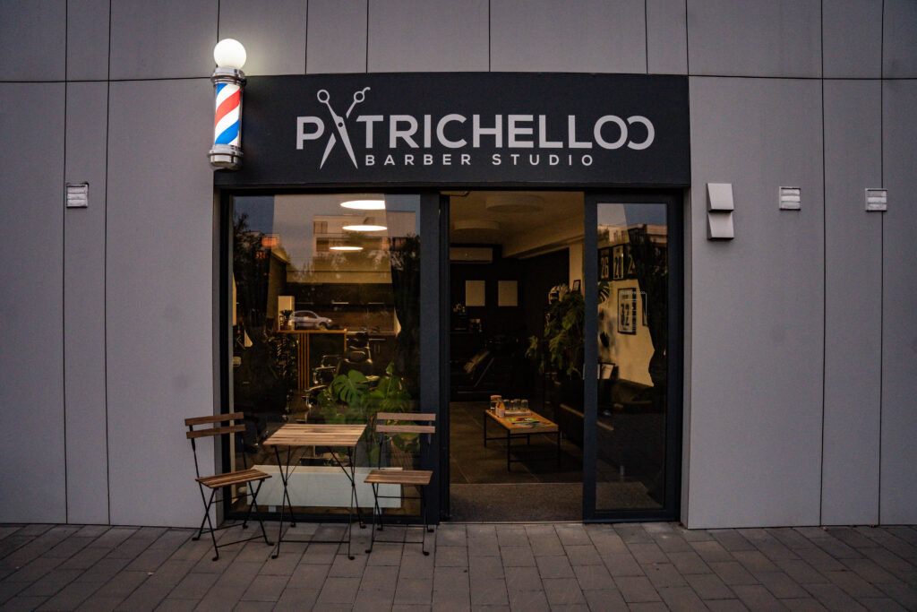 Fasáda Patrichelloo Barber Studio s osvětleným barber pole v podvečer